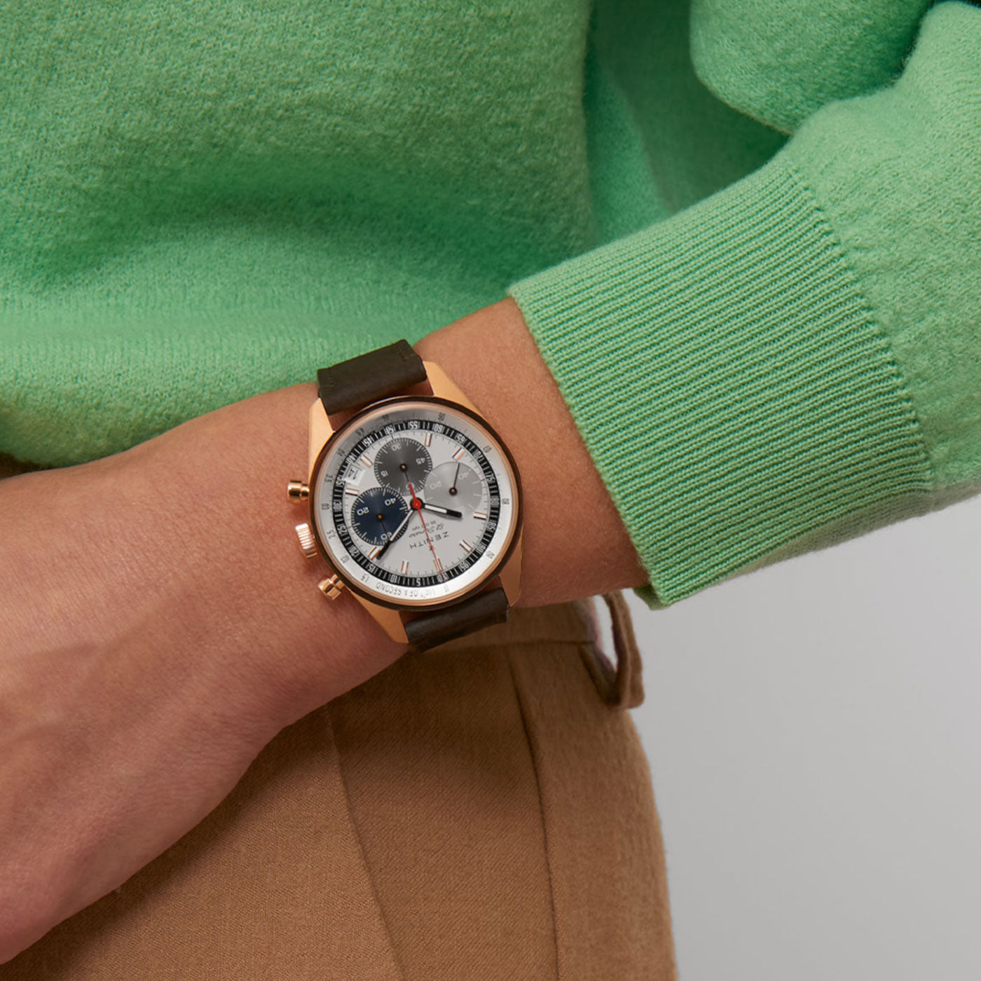 A person wearing the ZENITH Chronomaster Original El Primero 3600 Automatic Chronograph 38mm Watch by Zenith, featuring a gold case and brown leather strap, with a white dial adorned with black and red sub-dials. They pair this elegant timepiece with a green sweater and beige pants, creating an effortlessly sophisticated appearance.