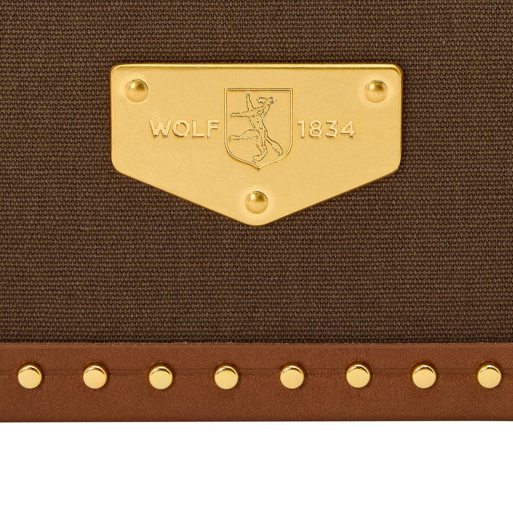 A close-up of the brown fabric surface of the WOLF 1834 Ida Trunk Jewellery Box showcases a gold emblem embellished with "WOLF 1834" and an engraved wolf image. Below, brass stud detailing accentuates the design, underscoring the luxurious essence of this limited edition 190th anniversary packaging by WOLF.