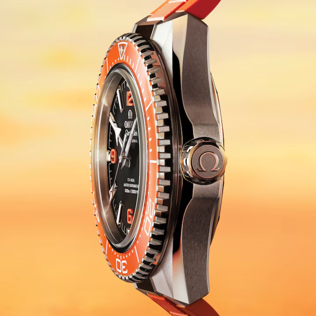 A close-up side view of the OMEGA Seamaster Planet Ocean 600M Co-Axial Master Chronometer 42mm watch with a black dial, orange bezel, and red strap, set against a warm orange-yellow gradient background.