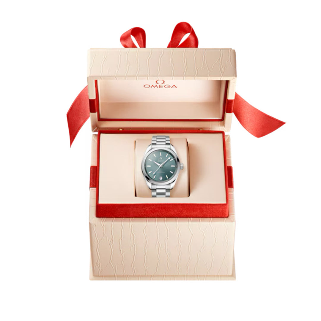 Inside an open, textured cream OMEGA box with red lining and a bow, lies the OMEGA Seamaster Aqua Terra Shades Co-Axial Master Chronometer 34mm Watch. It features a silver case, green face, and stainless steel bracelet.