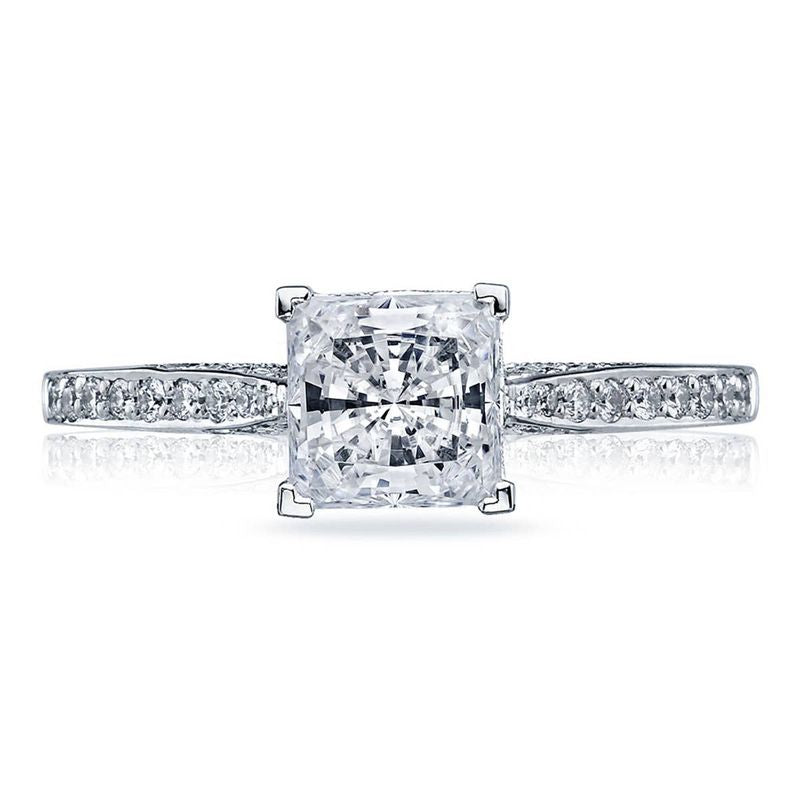 The Tacori Dantela Platinum Princess Cut Semi Mount Solitaire Engagement Ring with Diamond Band by Tacori showcases a stunning square diamond in the center, secured by four prongs. Its band is adorned with smaller diamonds, offering an elegant and reflective setting that enhances the brilliance of these exquisite gems.