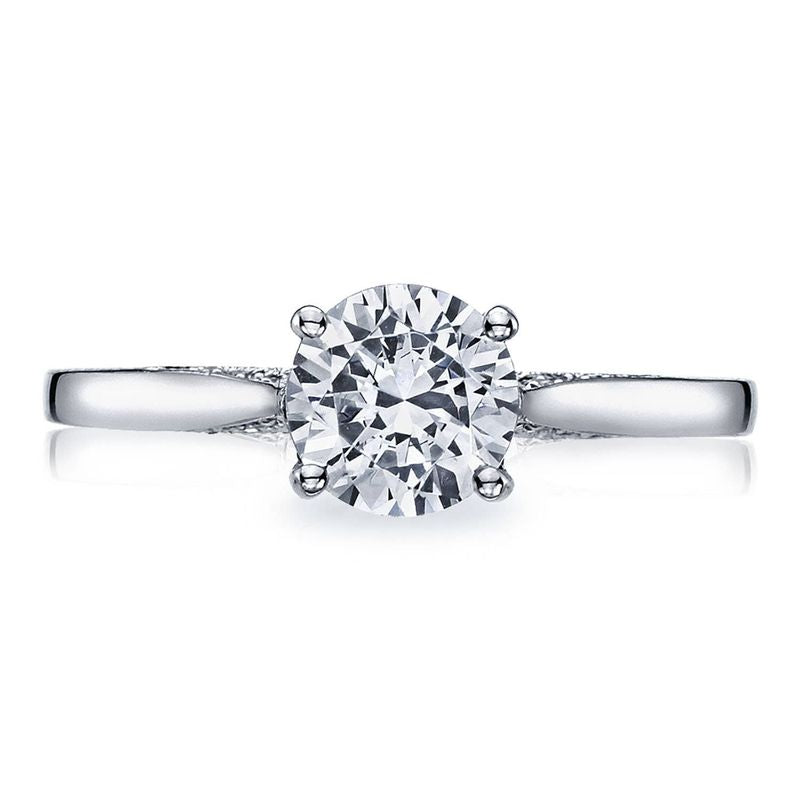 The Tacori Dantela 18K white gold round brilliant semi-mount solitaire engagement ring with a diamond band is displayed against a white background. The ring's dazzling solitaire diamond, expertly crafted by Tacori gemologists, serves as the focal point, reflecting light with an exquisite sparkle.