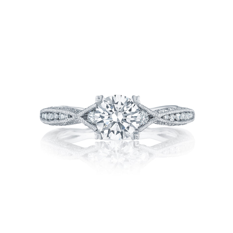 The Tacori Classic Crescent 18K White Gold Round Brilliant Semi Mount Solitaire Engagement Ring with Diamond Band showcases a central round diamond. The band is intricately designed with an intertwined pattern and adorned with smaller diamonds along the sides, set against a white background.