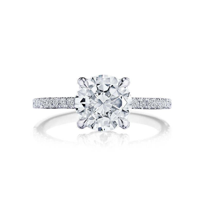 The Tacori Simply 18K White Gold Round Brilliant Semi Mount with Diamond Band and Hidden Halo Engagement Ring is beautifully highlighted in a close-up view. It features a prominent round diamond centerpiece, accompanied by pavé-set diamonds on the band that enhance its elegant and sparkling appearance. Expertly crafted by Tacori gemologists, this exquisite piece is set against a white background.