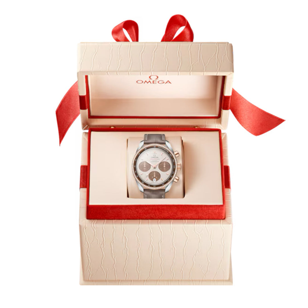 The OMEGA Speedmaster 38 Co-Axial Chronometer Chronograph 38mm Watch, displayed in an open cream-colored box with a red interior and ribbon, features a silver-tone case, dark dial with three subdials, and black strap. The prestigious Omega logo adorns the box lid.