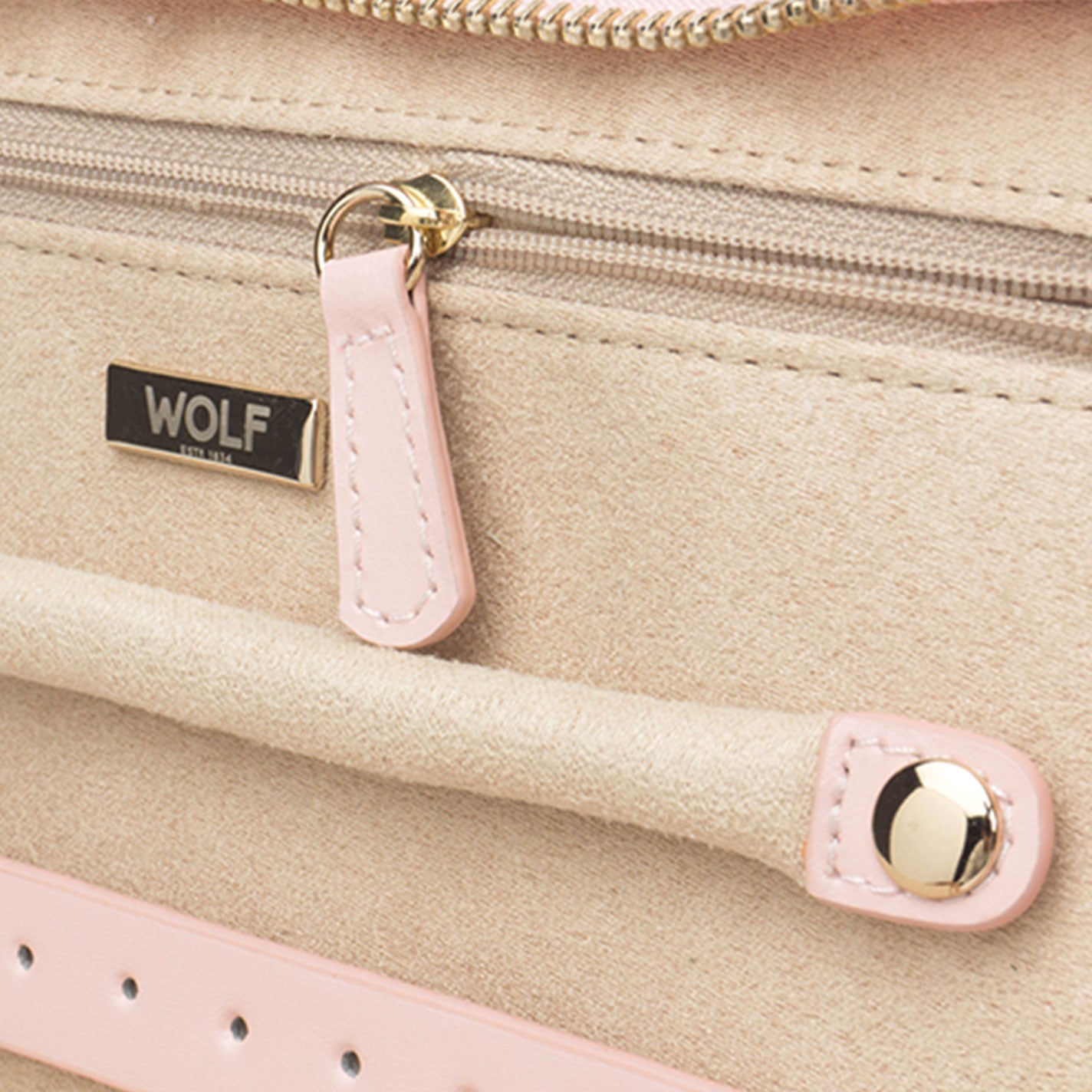 A close-up of the WOLF 1834 Caroline Pink Jewellery Box showcases its elegant design, featuring beige and pink diamond quilted leather with a small metal label reading "WOLF." The zipper includes a stylish pink pull tab, complemented by a gold button and pink trim on the handle, epitomizing sophistication and style.