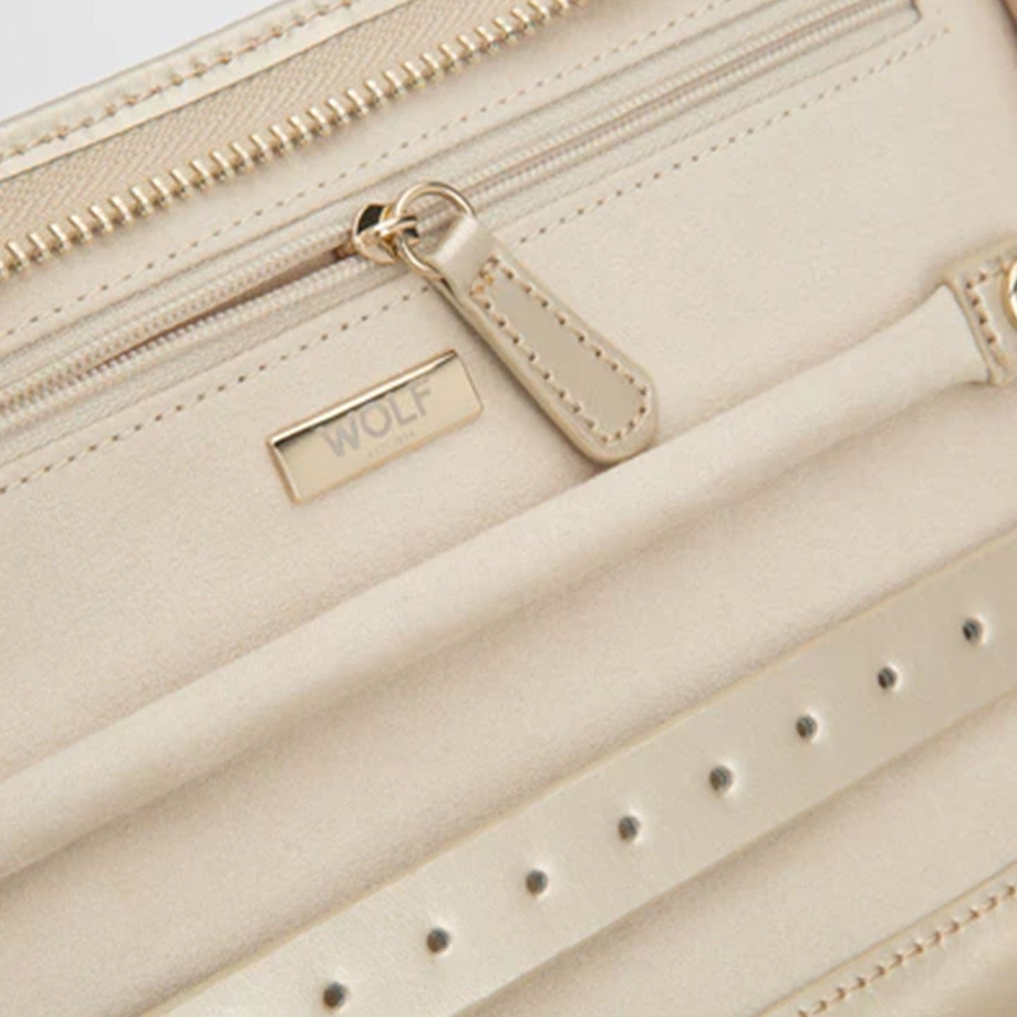 A detailed view of a cream-colored leather accessory showcases zipper and strap elements, enhanced by diamond quilted leather. Below the zipper lies a small metal plaque engraved with "WOLF," while the material exhibits a smooth, matte finish ideal for any WOLF 1834 Caroline Jewellery Portfolio.