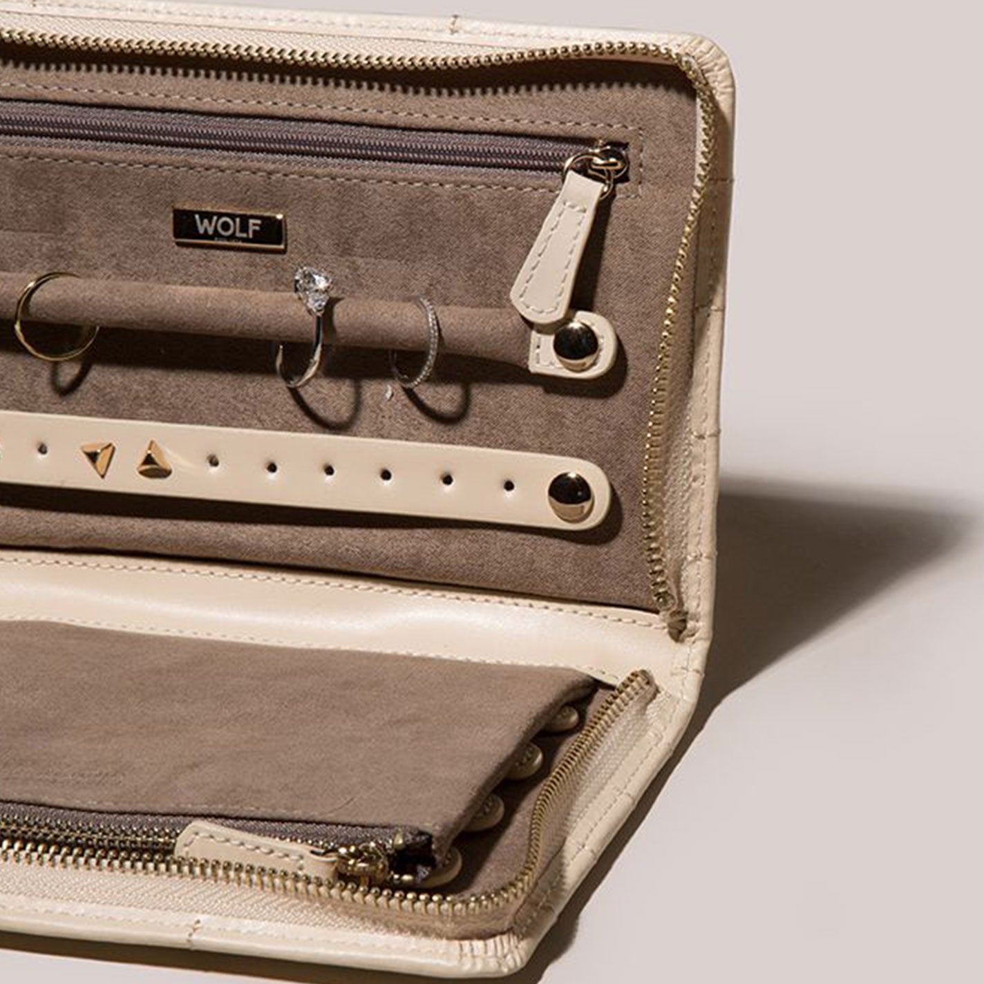 The WOLF 1834 Caroline Jewellery Portfolio, in beige leather with diamond quilted detailing, is open to display a diamond ring, silver hoop earrings, and triangular studs. It features the WOLF logo, a zipper closure, and LusterLocâ„¢ lining to protect your treasures.