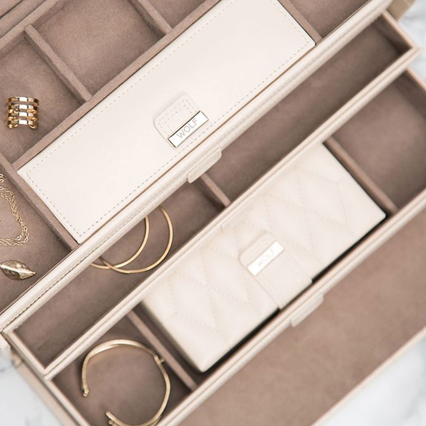 The WOLF 1834 Caroline White Large Jewellery Box exemplifies elegance with its inclusion of a beige clutch, gold bracelets, rings, and a necklace featuring a leaf pendant. Made from diamond quilted leather and lined with soft brown material, it offers ample storage within its compartments while LusterLocâ„¢ technology safeguards your treasures.