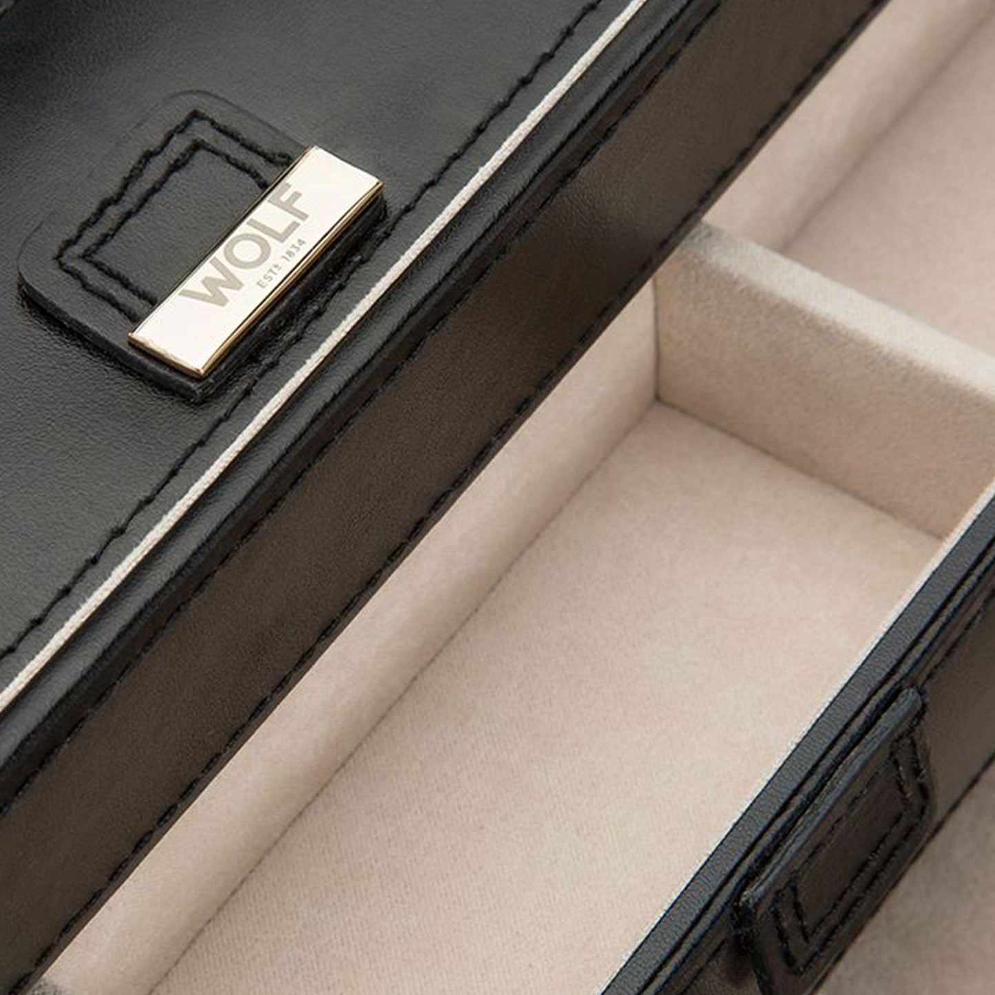 A close-up of the WOLF 1834 Caroline Black Large Jewellery Box, crafted with black diamond quilted leather and adorned with a gold-tone metal label featuring the brand name "WOLF." The interior is lined with soft beige fabric equipped with LusterLocâ„¢, providing compartments for watches or jewelry storage.