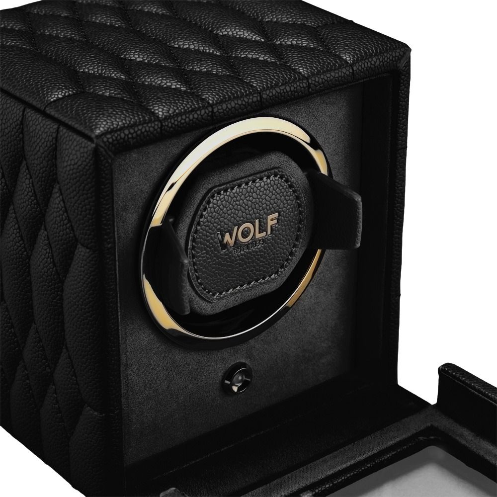A close-up of the WOLF 1834 Lauren Single Watch Winder Black showcases its black quilted leather exterior with gold accents and chrome-finished hardware, featuring the WOLF logo on the padded holder. The open box reveals an ebony macassar interior and a secure watch holder inside.