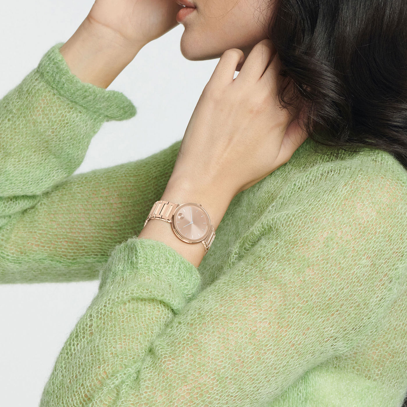 A person with long, dark hair adjusts their hair while wearing the Movado BOLD Horizon Quartz 34mm Watch in rose gold. They are dressed in a fuzzy green sweater, with the focus on the watch and the textured fabric of the sweater.