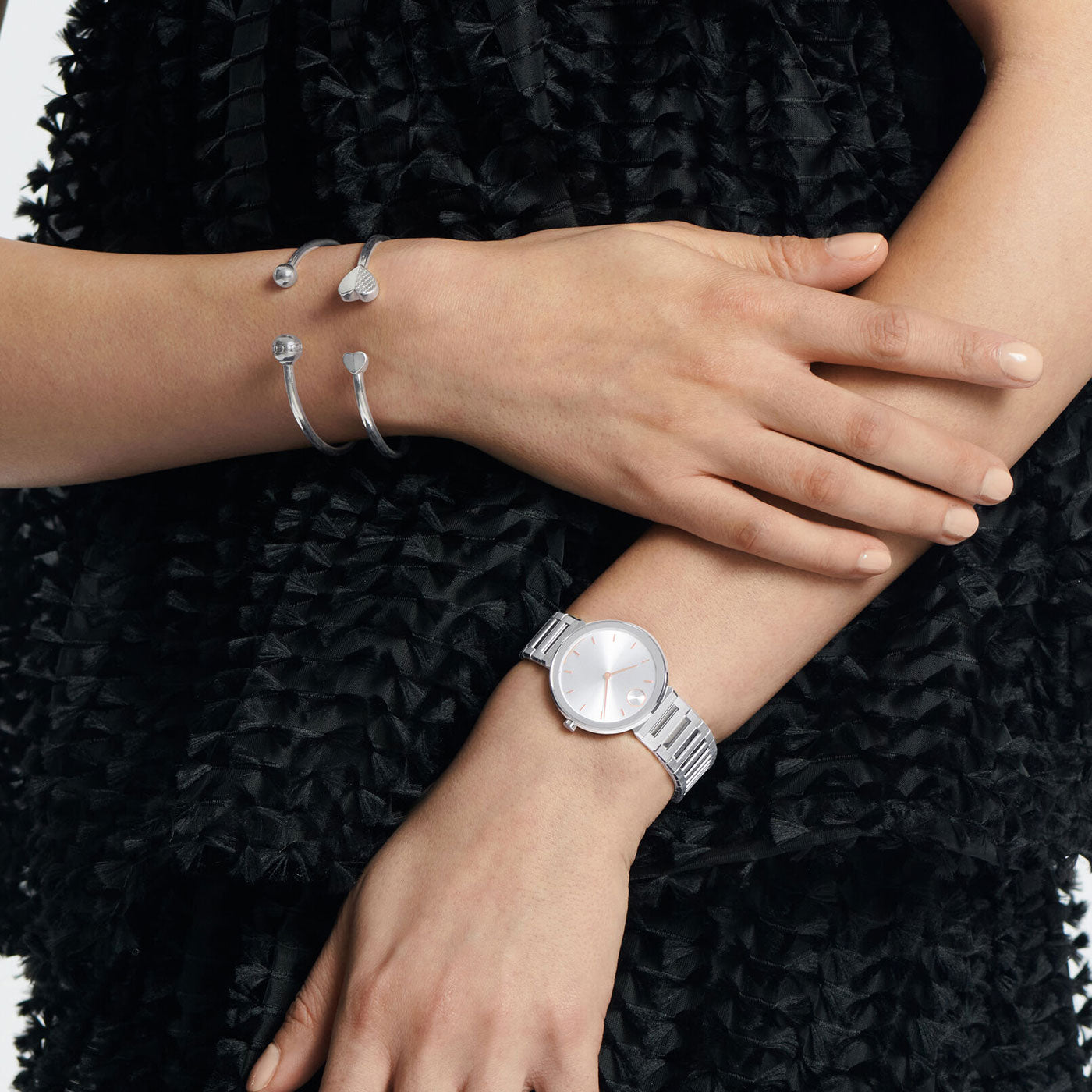 A person in a black textured dress crosses their arms, showcasing the elegant Movado BOLD Horizon Quartz 34mm Watch and two stylish silver bangles on one wrist. The watch features a simple round face and a metal link band.