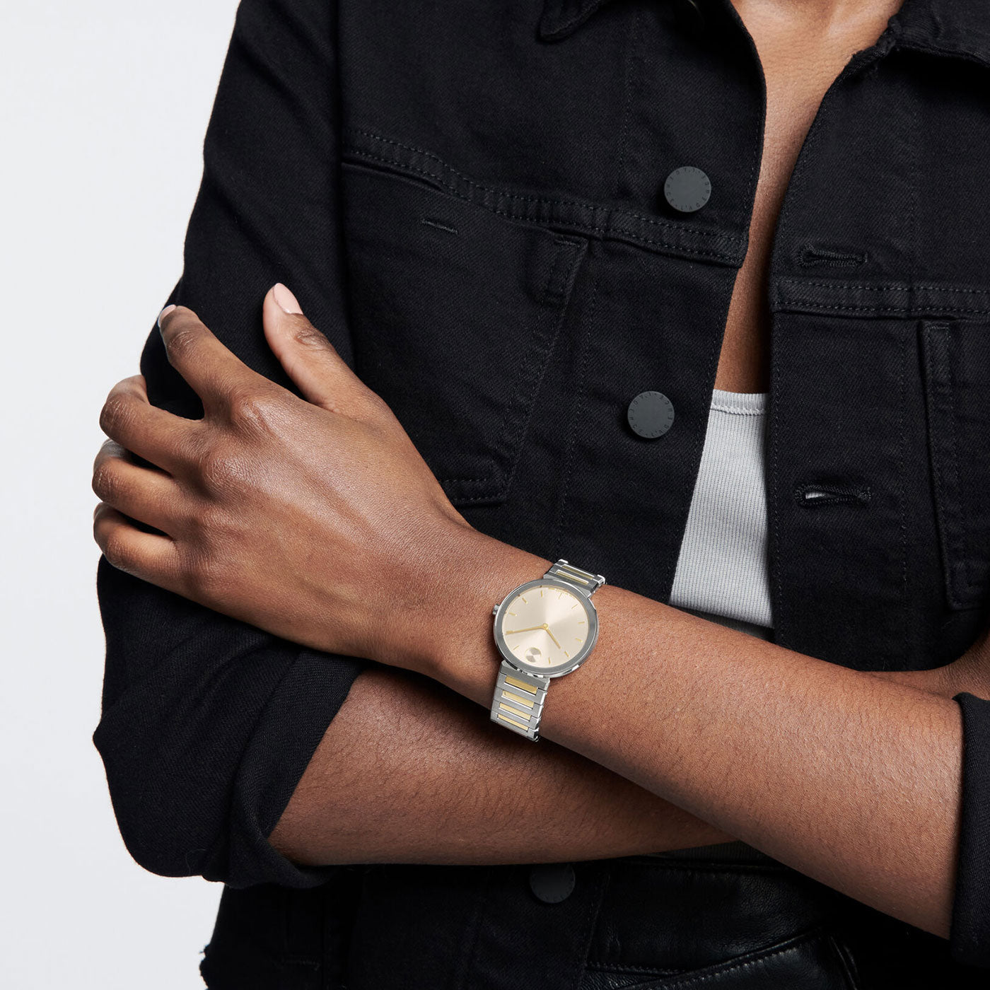 Person wearing a black denim jacket and a light-colored top, with arms crossed. They have a Movado BOLD Horizon Quartz 34mm Watch featuring a metallic strap on their left wrist.