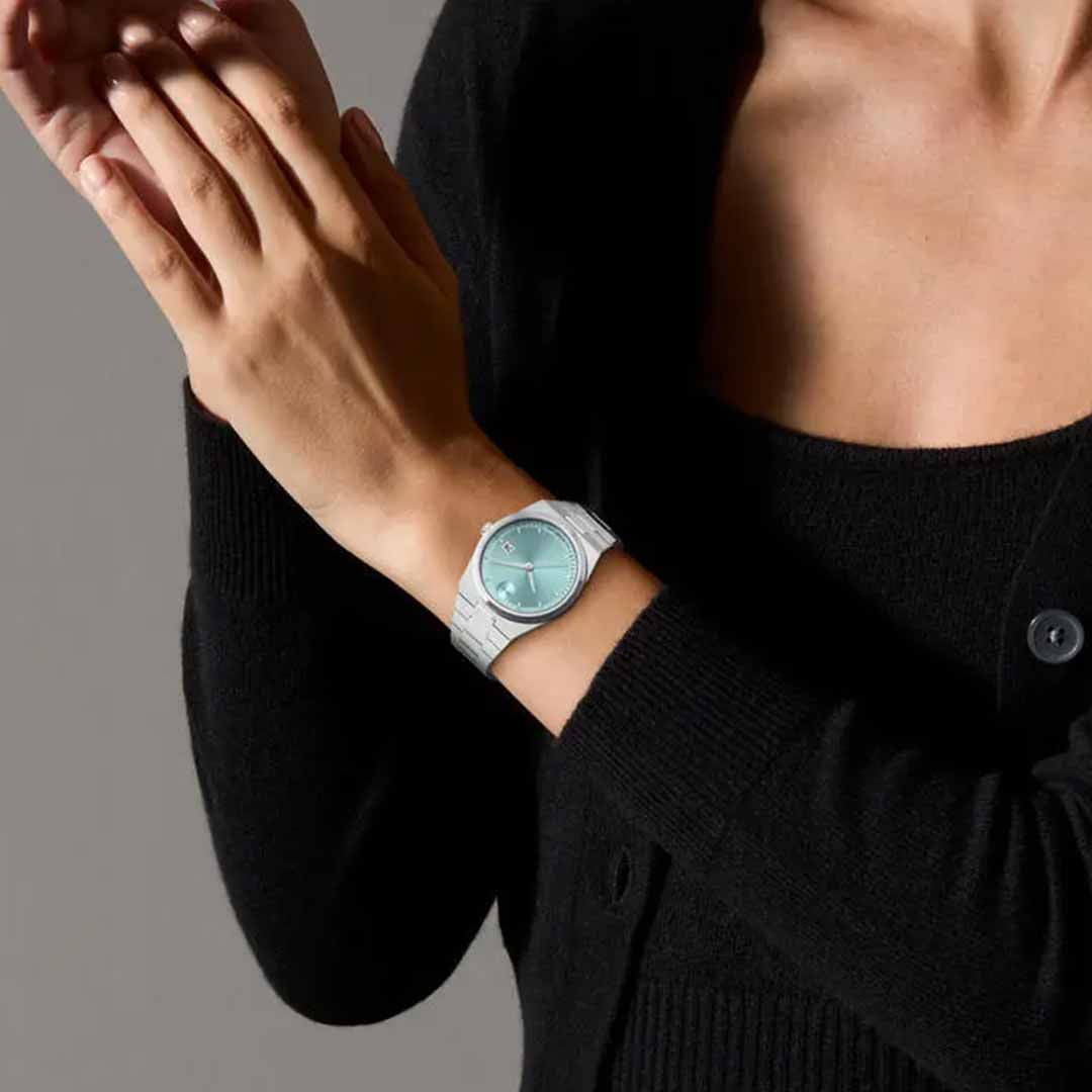 Wearing a black sweater, someone adjusts the Movado BOLD Quest Quartz 35mm Watch, which showcases stainless steel hands and a seafoam blue sunray dial with a white band, set against a neutral gray background.