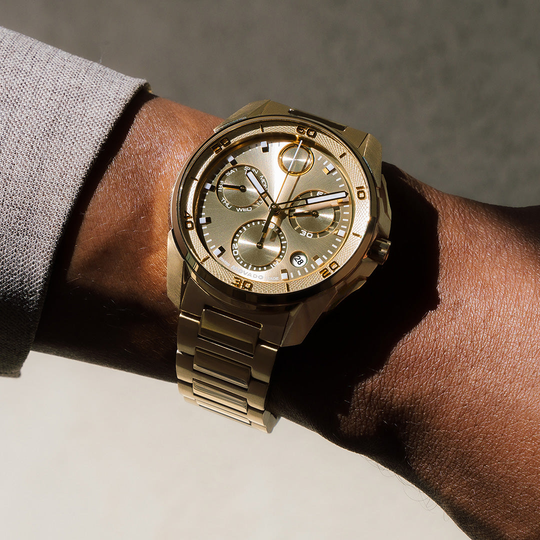 Movado BOLD Verso Quartz Chronograph 44mm watch in gold-tone with a textured dial, three subdials, and a metal link bracelet worn on a wrist.
