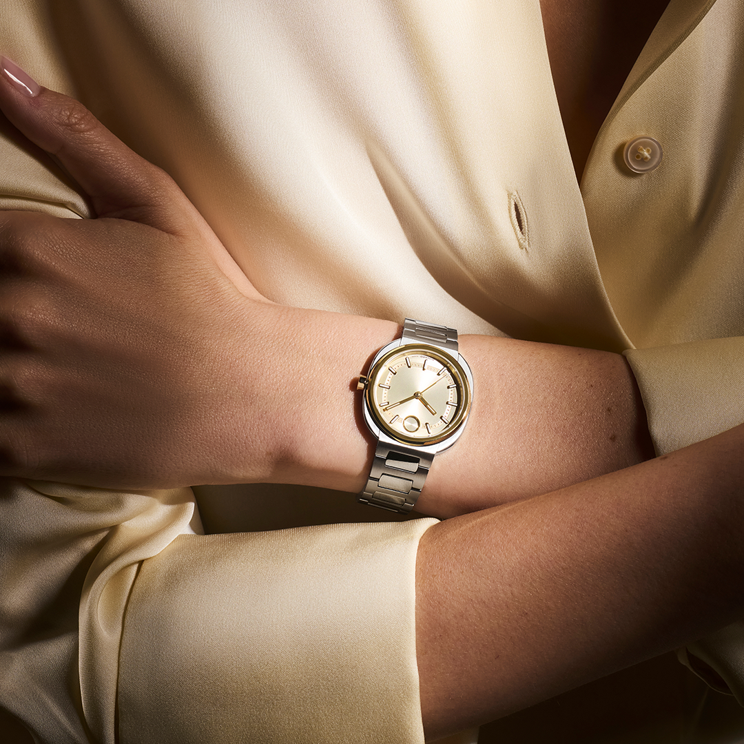 A Movado BOLD Nuvo Quartz 30mm X 31mm Watch featuring a round, yellow gold ion-plated bezel and a silver-toned metal link bracelet worn on a wrist.