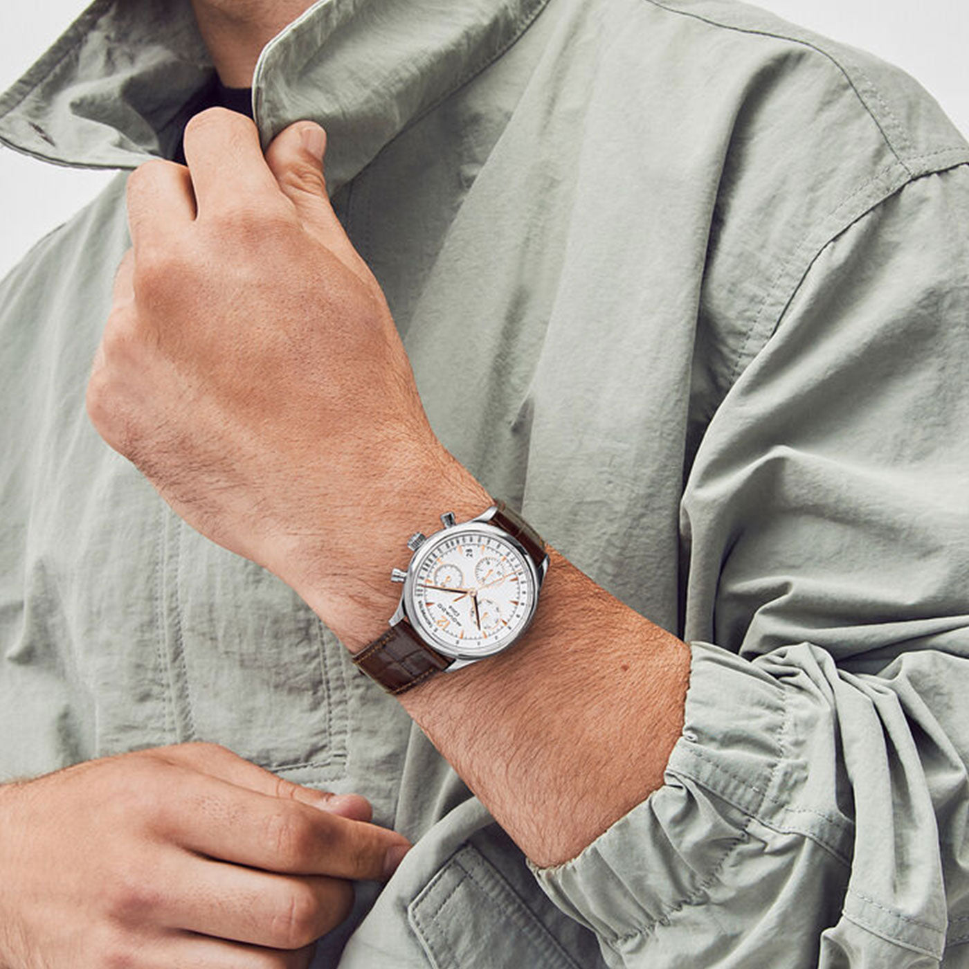 A person wearing a light gray jacket is adjusting their cuff, revealing a Movado Heritage Series Circa Solar 43mm watch. The timepiece features a stainless steel case and a brown leather strap. Its striking chronograph dial with white and silver accents beautifully complements the ensemble.