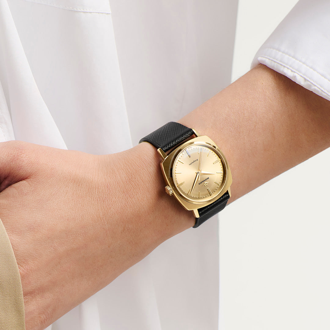A gold-tone Movado Heritage Kingmatic Automatic 35mm Watch with a round champagne-colored dial and a black textured strap worn on a wrist.