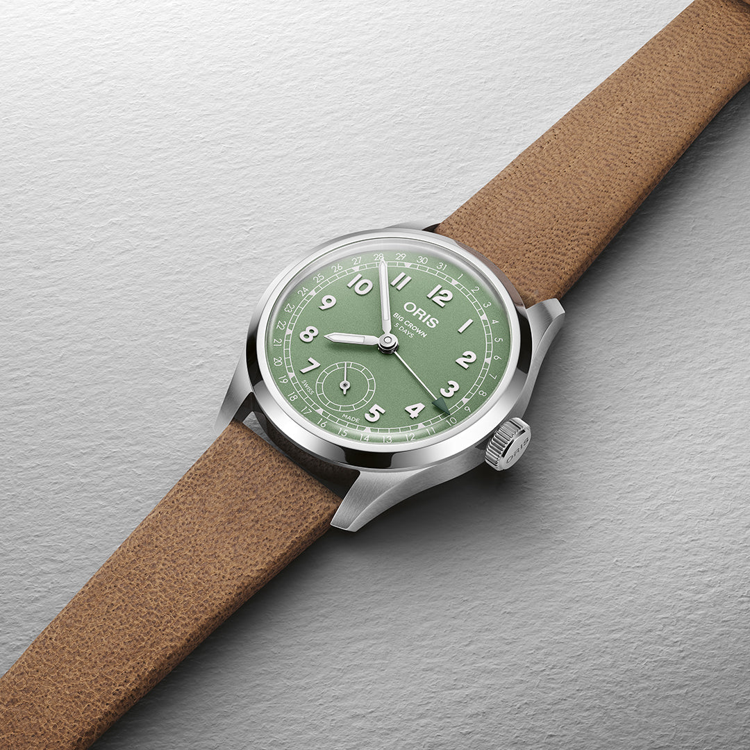 The ORIS Big Crown Pointer Date Calibre 403 features a green face with white numerals, a subdial, and is complemented by the timeless Calibre 403. It boasts a silver casing and textured brown leather strap against a textured gray backdrop for an exquisite display.