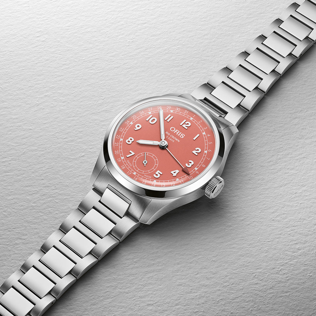 The ORIS Oris Big Crown Pointer Date Calibre 403 Automatic Winding 40mm Watch features a coral dial with white numerals, a second sub-dial, and is powered by Calibre 403 automatic movement. Housed in stainless steel, its link bracelet elegantly sits at an angle on textured light gray.
