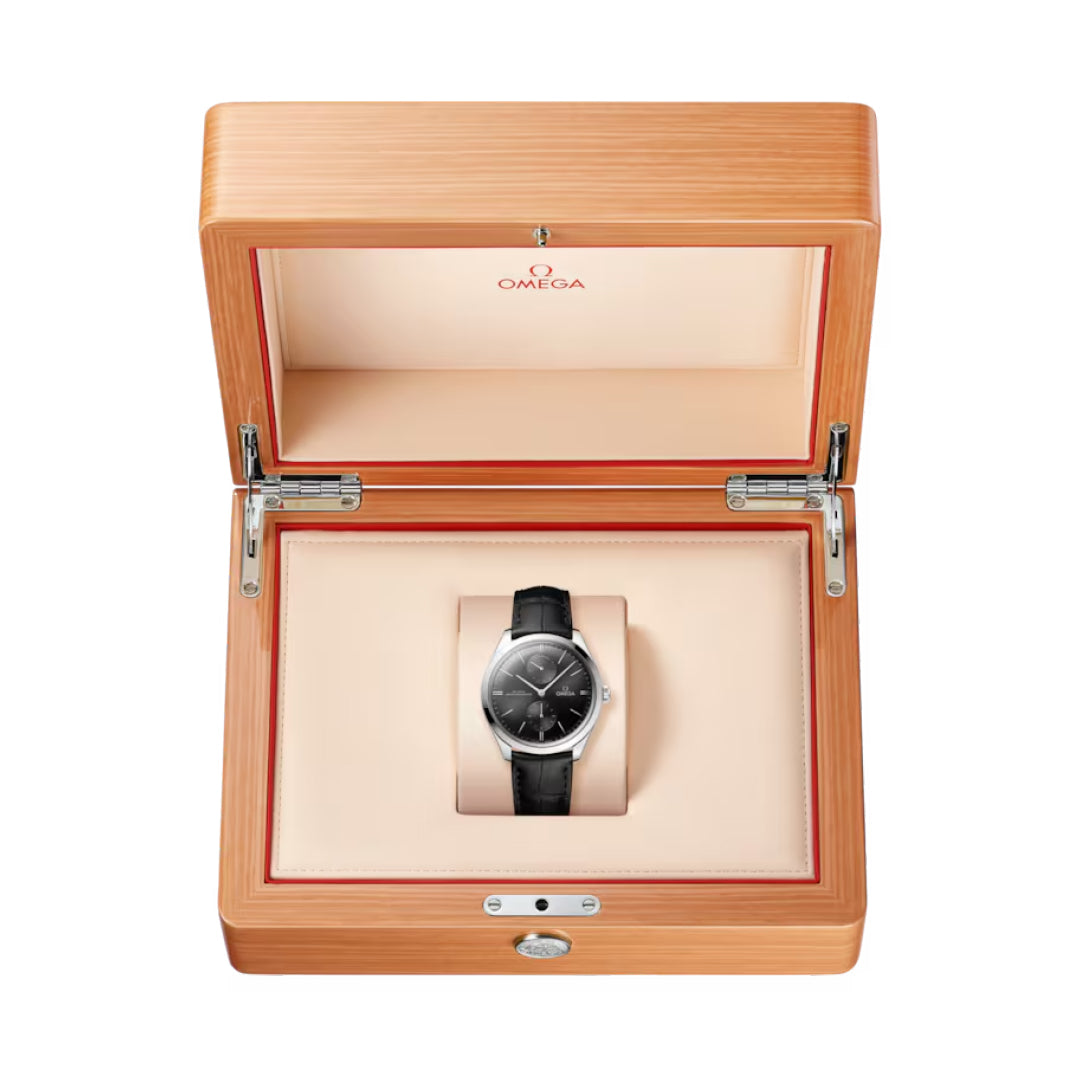 The OMEGA De Ville Tresor Co-Axial Master Chronometer 40mm watch, featuring a black leather strap, dark dial, and 18K white gold case, is showcased in an open wooden Omega box lined with cream padding.