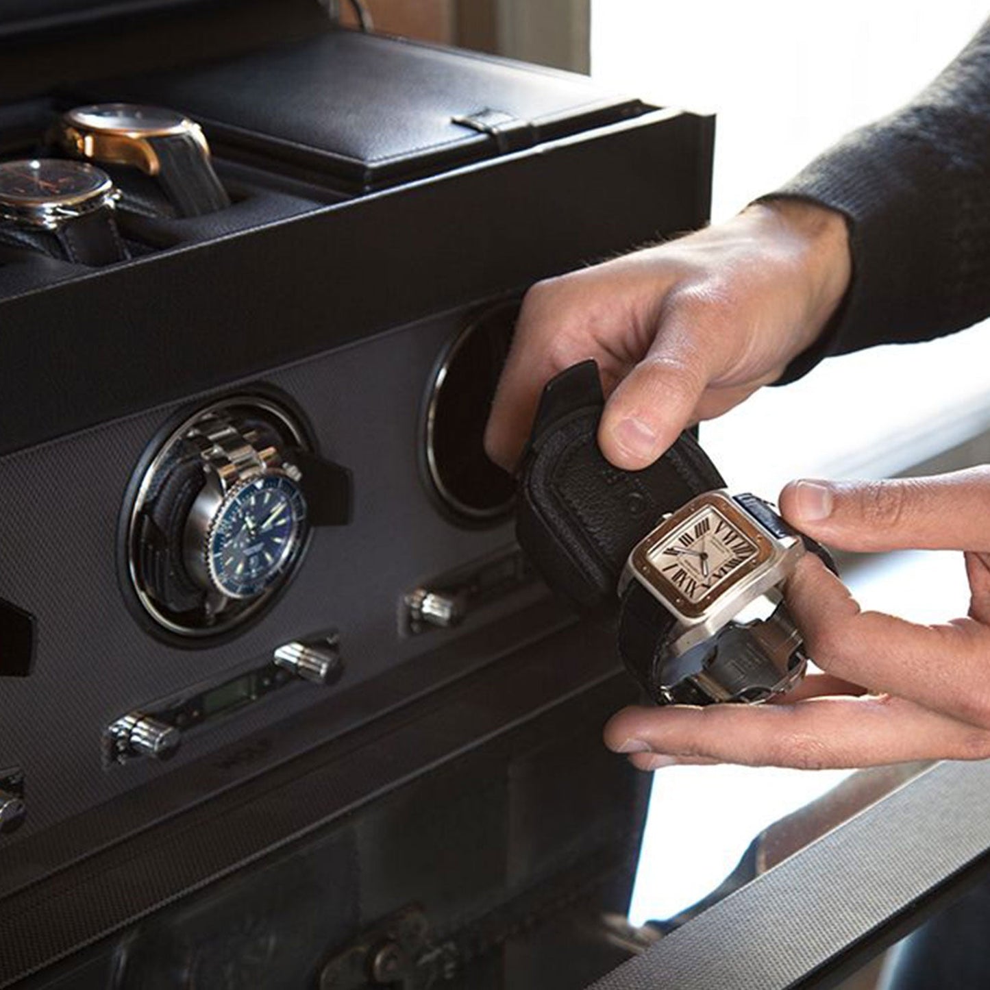 A person is holding a square-faced watch with a leather strap. In the background, the WOLF 1834 Viceroy Triple Watch Winder with Storage elegantly displays several other watches. This sleek black winder by WOLF is renowned for its patented innovation and various rotation options, and it features a glass cover.
