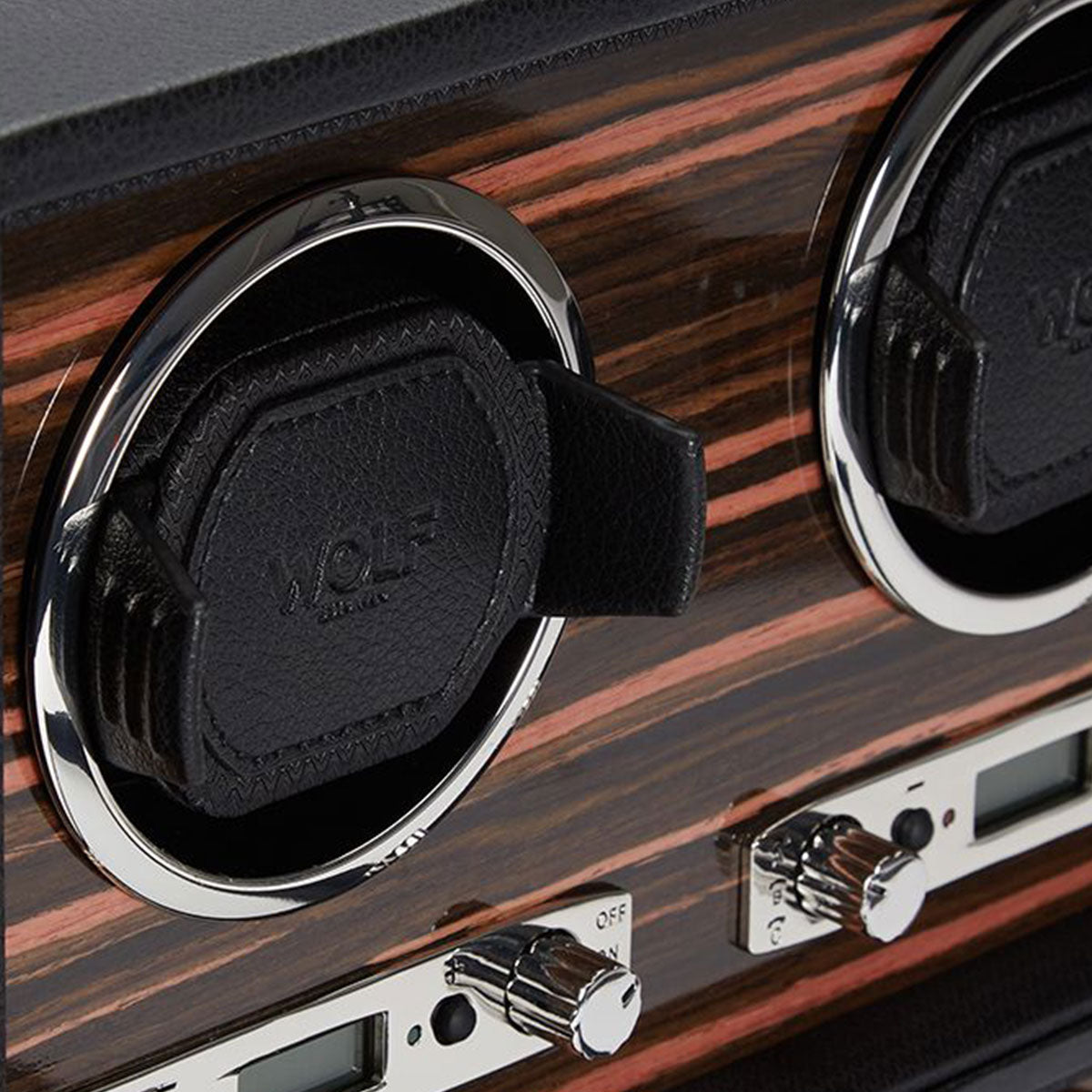A detailed view of the WOLF 1834 Roadster 4 Piece Winder showcases its Ebony Macassar design, featuring a dark wood finish complemented by silver accents. The interior reveals two black leather watch holders prominently labeled "WOLF." Beneath them are control knobs and switches that allow for setting adjustments, seamlessly merging elegance with functionality for any roadster aficionado.