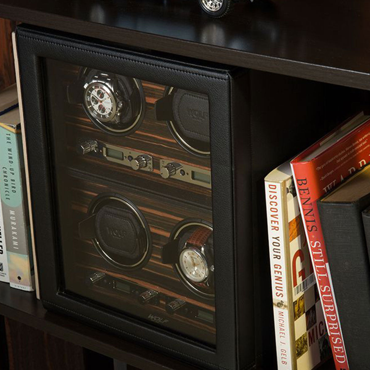 A WOLF 1834 Roadster 4 Piece Winder, beautifully crafted with an Ebony Macassar finish, elegantly displays two watches within its leather case and glass front, nestled on a bookshelf amidst a collection of books.