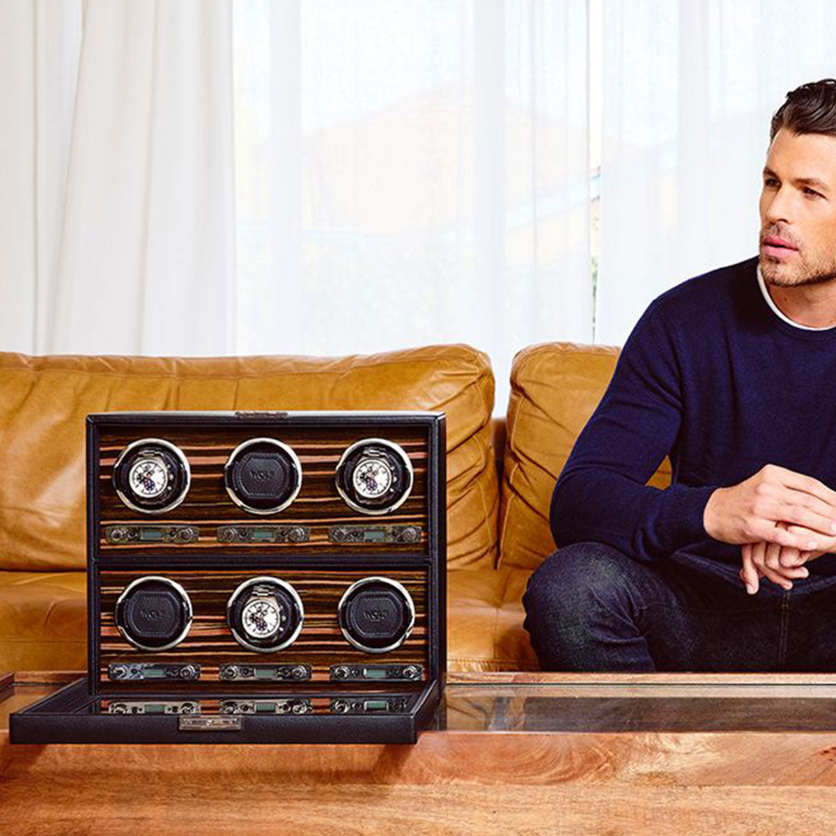 A man in a navy sweater sits on a tan leather sofa. Beside him on an ebony macassar table is an open case showcasing six luxury watches, featuring the WOLF 1834 Roadster 6 Piece Winder by WOLF. White curtains hang in the background.