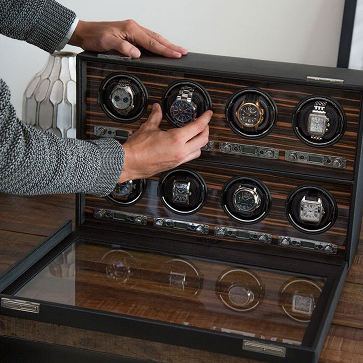 A person adjusts a wristwatch inside the WOLF 1834 Roadster 8 Piece Winder, which offers multiple rotation options and is placed on a wooden table. The winder features a glass cover and can hold several watches in circular holders. A decorative lamp softly illuminates the background, enhancing the elegance of the scene.