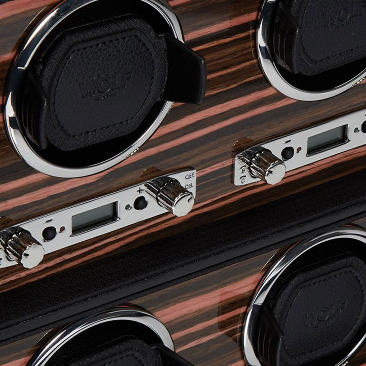 Close-up of the WOLF 1834 Roadster 8 Piece Winder, showcasing its luxurious dark wood finish. It features multiple circular slots with padded holders bearing the "WOLF" brand name, along with digital displays and metallic control dials for selecting rotation options. This watch winder offers precise power reserve settings.