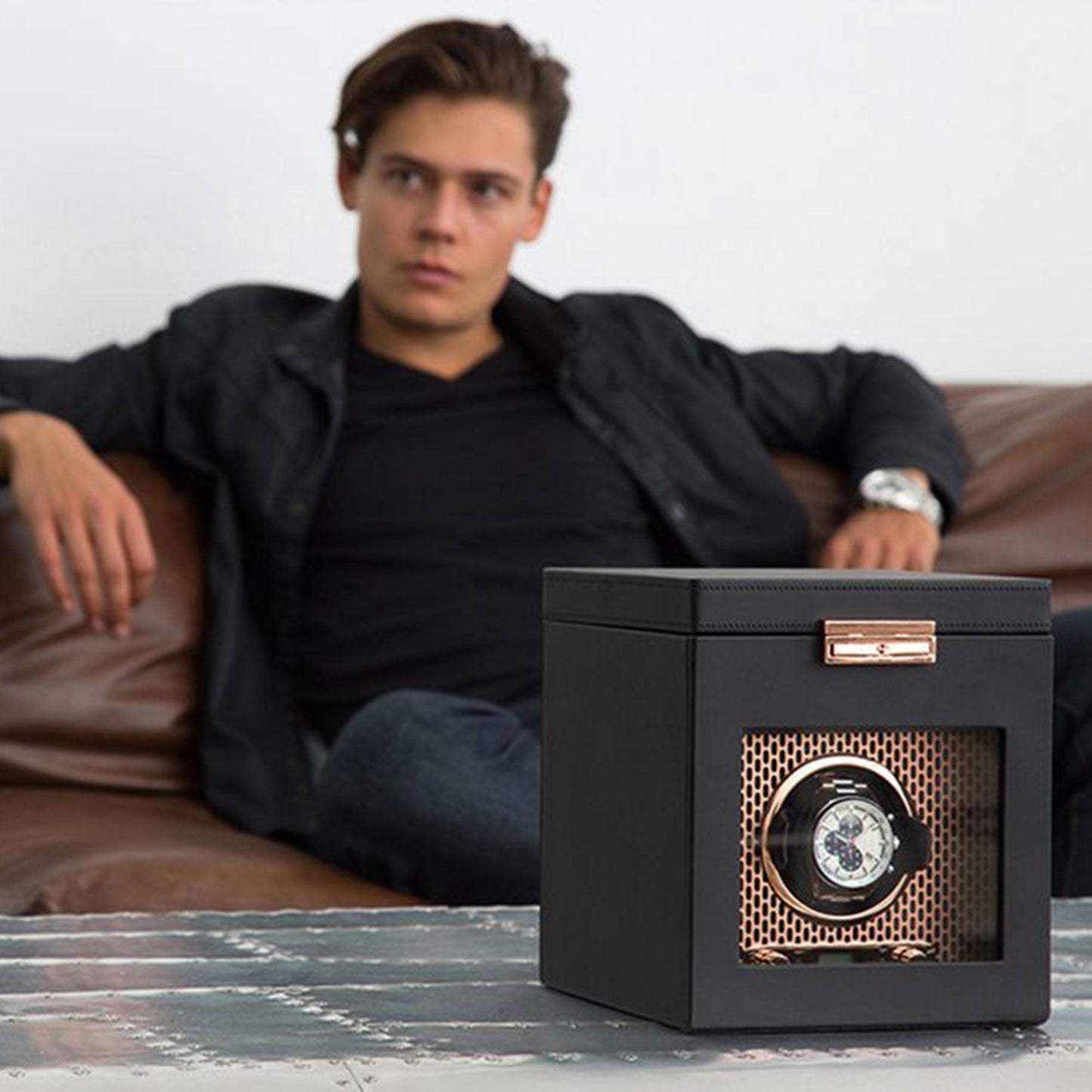 In the foreground, placed on a metal table, is the WOLF 1834 Axis Watch Winder by WOLF, featuring rotation options and a transparent front that beautifully showcases a timepiece. Meanwhile, in the background, someone relaxes on a brown leather couch.