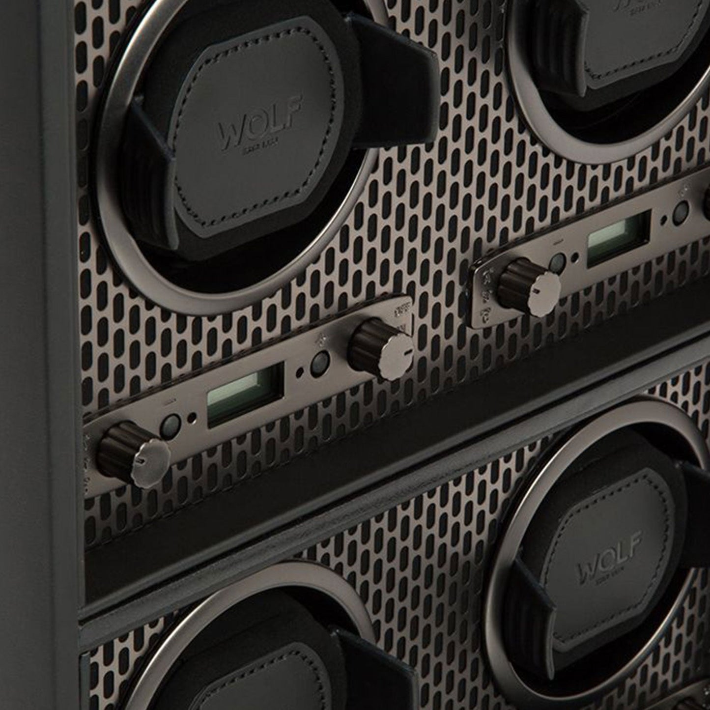 A close-up of the WOLF 1834 Axis 4 Piece Winder reveals a luxury timepiece storage solution with four compartments. Each section is equipped with a black leather pad, digital display, and dials for rotation options. The metal panel showcases a perforated design, and the brand name "WOLF" is prominently visible on the pads.