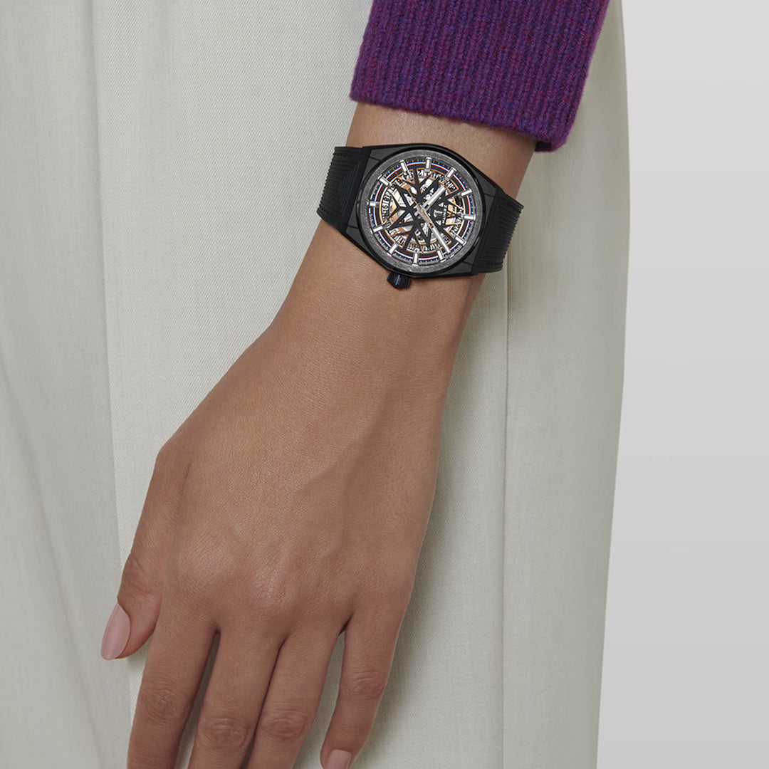 A person wearing a purple sleeve showcases a ZENITH Defy Classic Elite 670 SK 41mm watch with its intricate mechanical design and sleek black ceramic strap against a plain light background.
