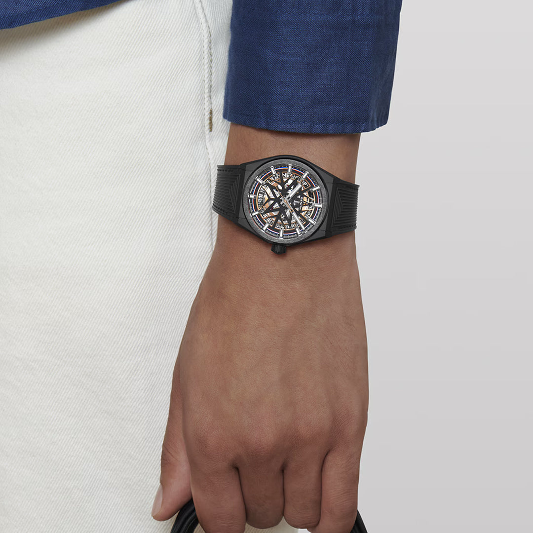A person wearing a blue jacket and white pants shows off a ZENITH Defy Classic Elite 670 SK 41mm watch, featuring a skeleton face and black ceramic band. The hand is relaxed with fingers gently touching a dark object, set against a neutral background.