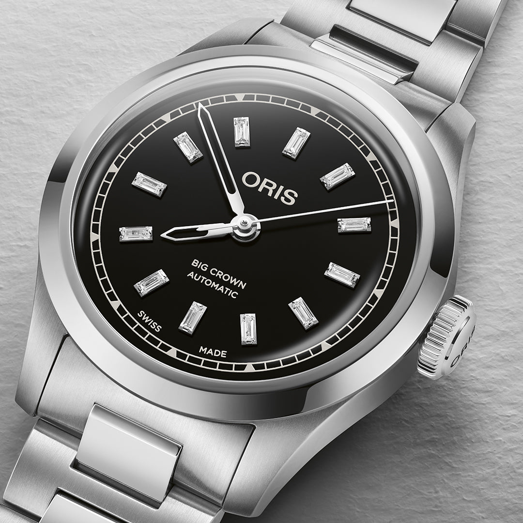 A close-up of the Oris Big Crown Calibre 531 Automatic Winding 34mm Watch showcases its elegance with a stainless steel band and silver case. The black dial features diamond hour markers, "Big Crown Automatic" and "Swiss Made," all resting on a textured gray surface.