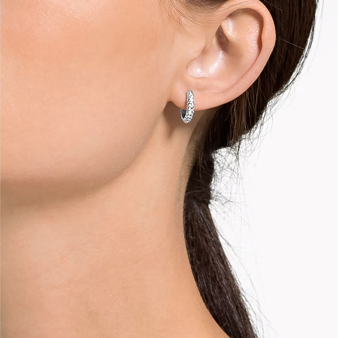 A close-up of a woman's ear showcases the sleek elegance of minimalist jewelry—a small hoop earring, the Swarovski Dextera Hoop Earrings, embellished with clear pavé stones. Set against a plain white background, the rhodium-plated design by Swarovski highlights both the contours of her ear and her neatly tied-back hair.