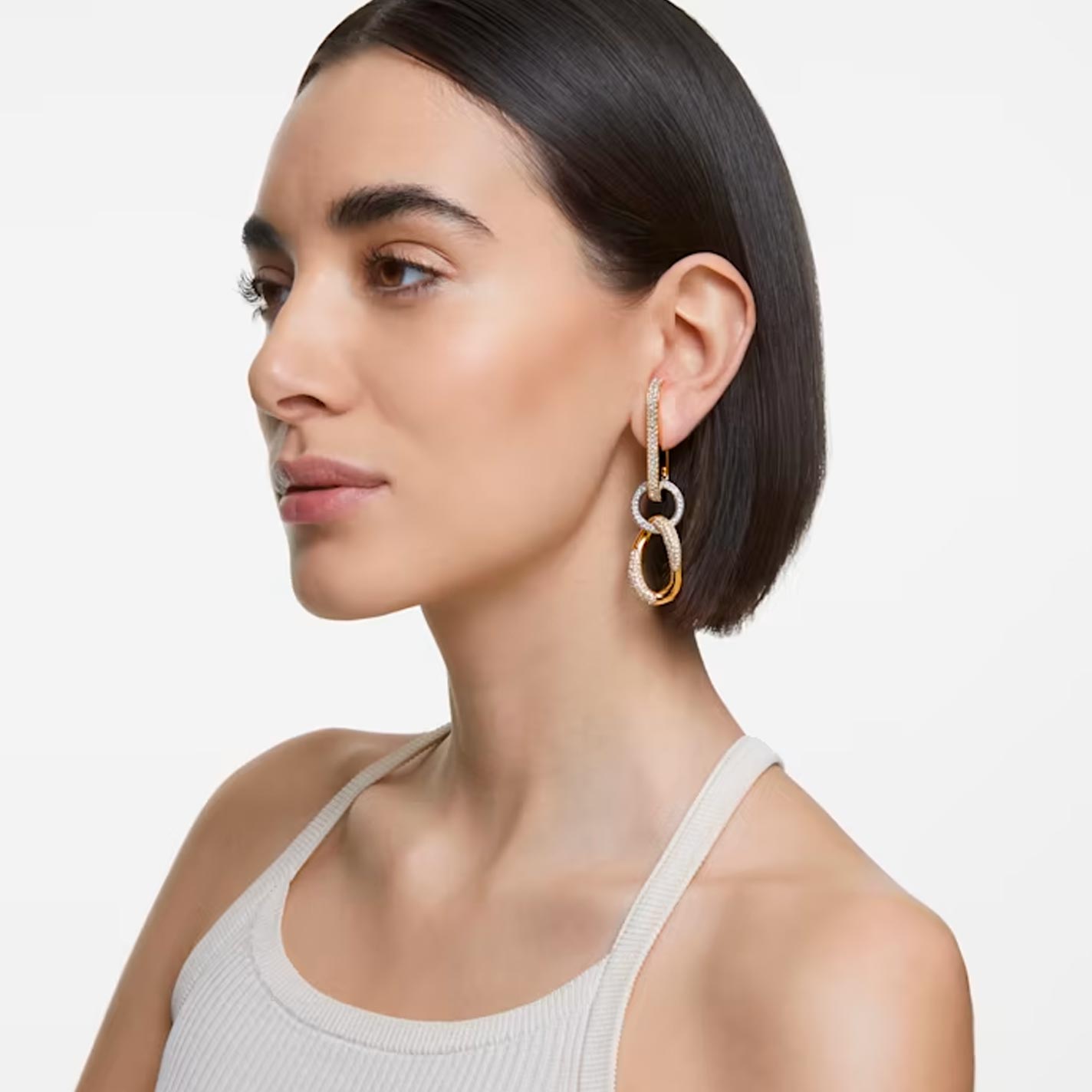 A person with short dark hair is wearing a white sleeveless top and Swarovski Dextera Chain Link Drop Earrings, which sparkle like clear crystal. They gaze sideways against a plain background, embodying the unique craftsmanship of Swarovski.