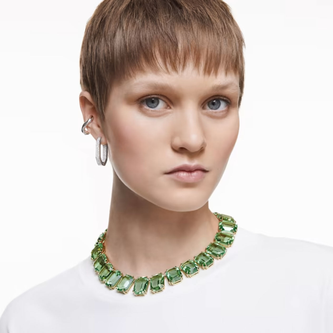 The individual with short, light brown hair dons a white shirt and displays an eye-catching necklace adorned with large green gems. Accompanying the ensemble is a pair of Swarovski Dextera Hoop Earrings, which adds elegance against the plain white background.