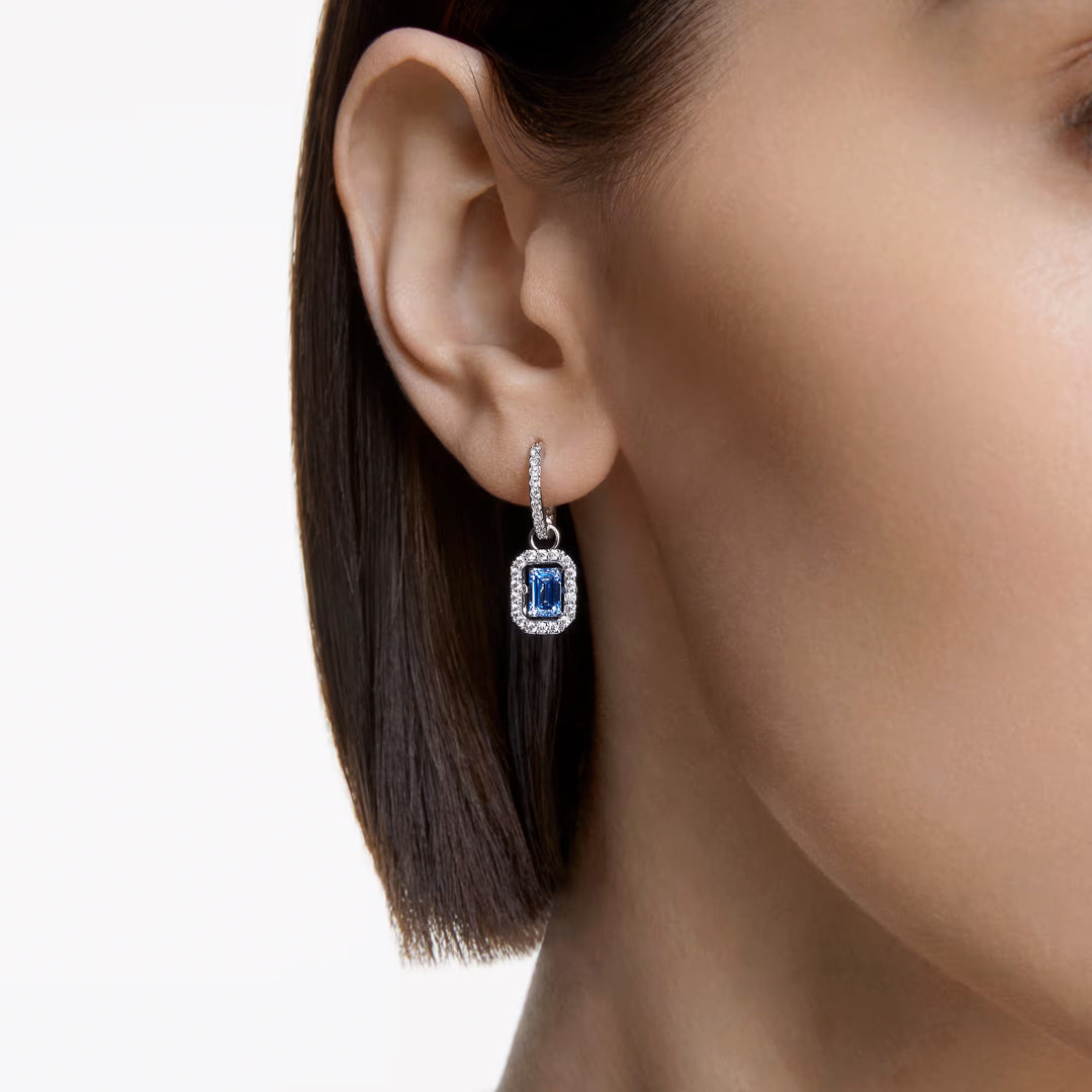 Close-up of a woman's ear adorned with the Swarovski Una Drop Earrings. The dangling design features a captivating blue dancing stone gracefully encircled by white pavé crystals in a silver or white gold frame. The woman’s short, straight hair perfectly complements the elegant sparkle of these Swarovski earrings.