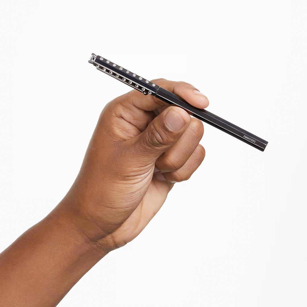 A close-up shot showcases a hand elegantly holding the Swarovski Ballpoint Pen, featuring silver details and adorned with crystals from the Millenia Family, set against a white background.