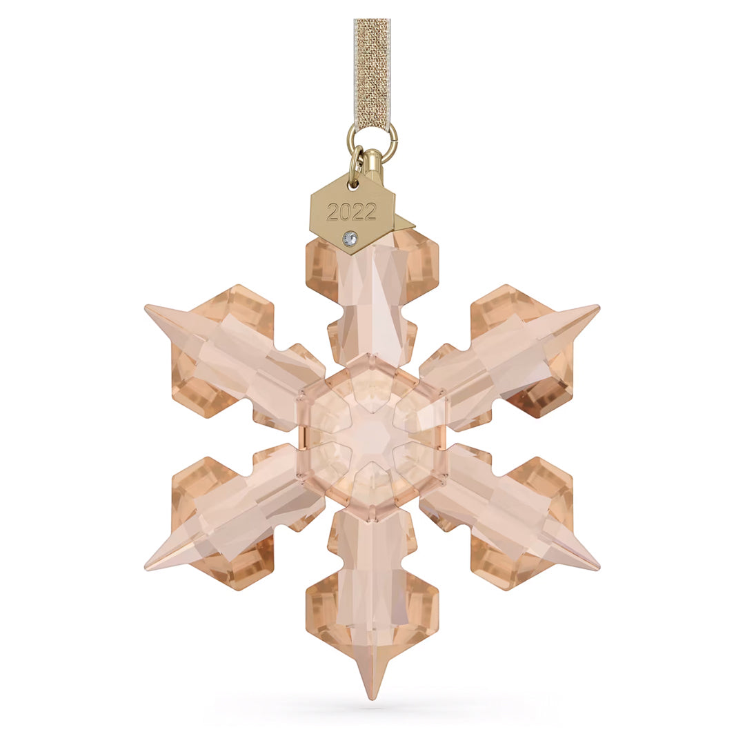 A Swarovski Festive2022 Annual Edition Ornament Home Décor piece elegantly adorns a space with its light amber-hued crystal snowflake design, hanging from a gold ribbon. It showcases intricate facets surrounding a hexagonal center and is adorned with a small gold tag engraved "2022" and a gemstone accent, making it perfect for holiday decorations.