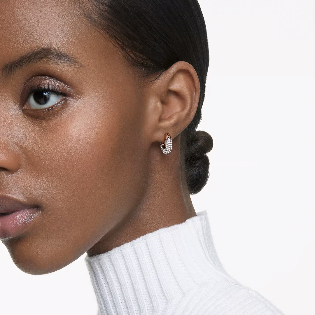 A close-up captures a person with a tidy hairstyle wearing Swarovski Dextera Hoop Earrings in a rose gold tone. They are dressed in a white, ribbed turtleneck sweater set against a plain backdrop. The image emphasizes their left side, showcasing the earring prominently.