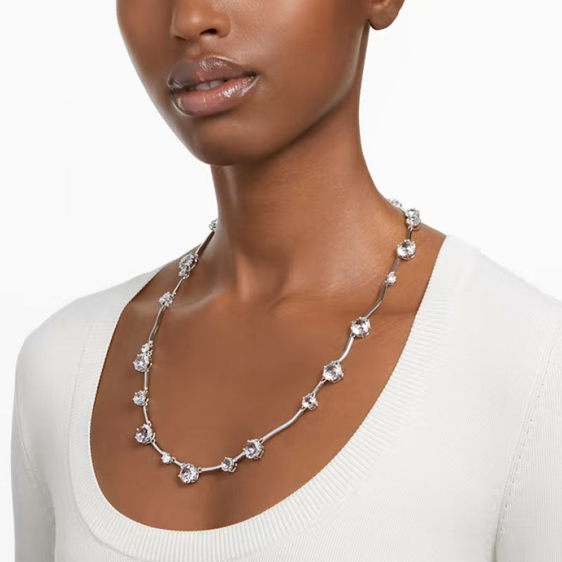 A person wearing a white top and a Swarovski Mixed Round Cuts Crystal Constella Necklace, featuring circular, clear gemstones that evoke the beauty of constellations. The rhodium-plated necklace rests elegantly along the neckline.