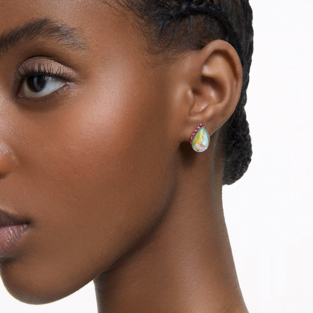 Close-up of a person wearing the Swarovski Chroma Stud Earrings by Swarovski, featuring a pear-shaped design plated in rhodium and embellished with crystals, all set in an eye-catching red border. The simple background highlights the earring's brilliance and perfectly complements the person's braided hair.