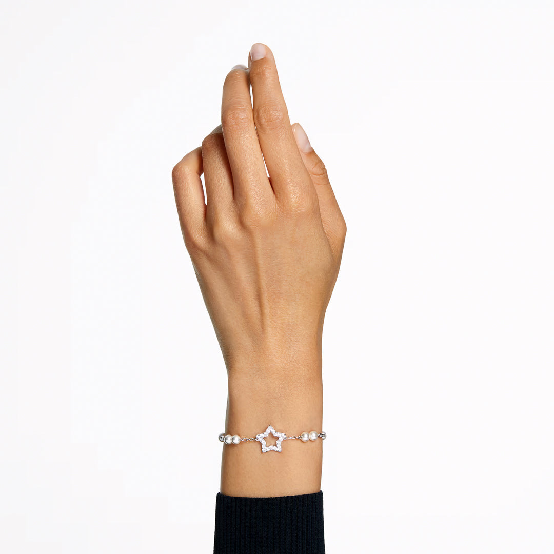 A hand wearing the Swarovski Stella Bracelet, featuring a rhodium-plated design with crystal pearls and a pink pavé-embellished star charm, is shown against a white background. The person is dressed in a black long-sleeve top.