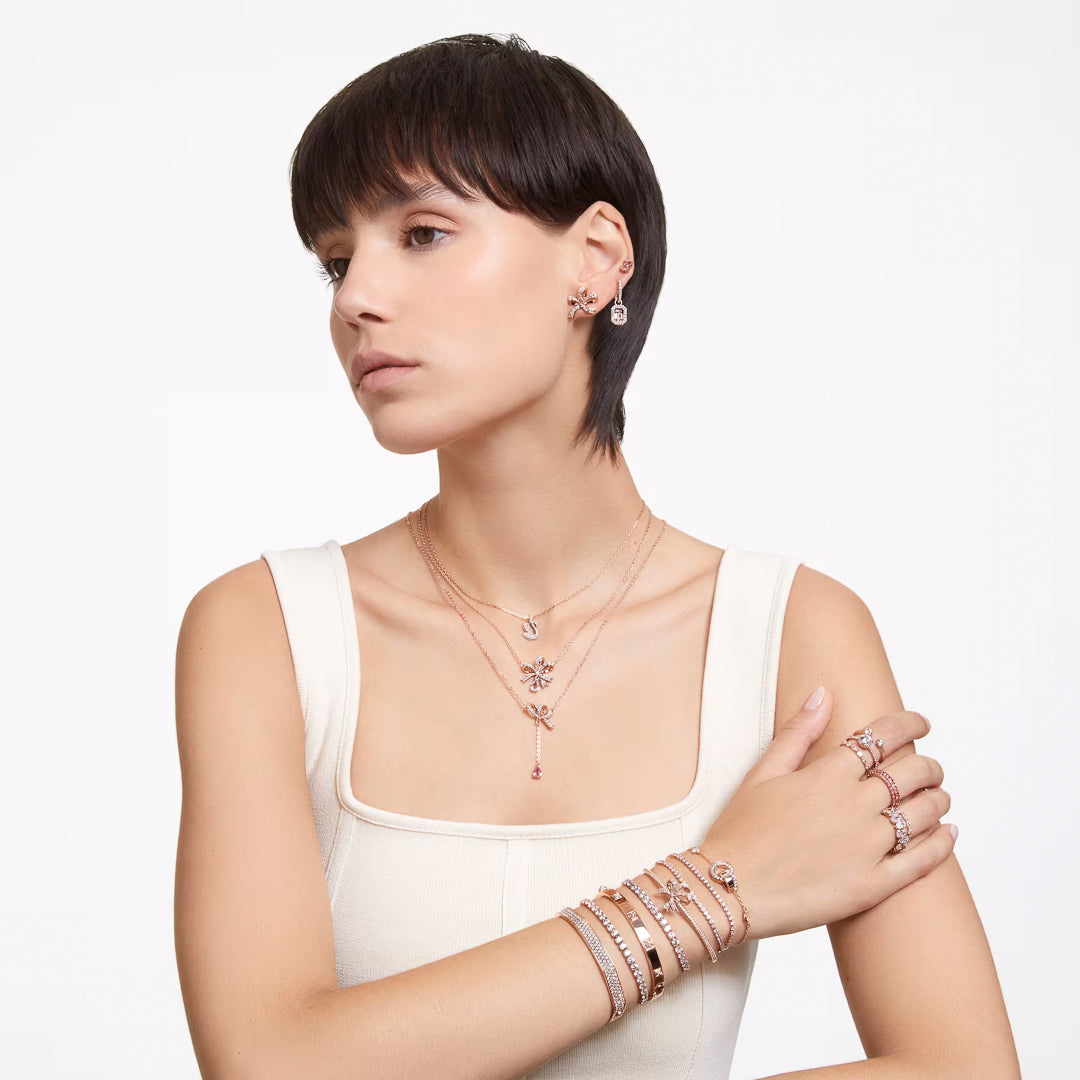 A woman with short brown hair is adorned in Swarovski jewelry, including rose gold-tone plated pieces with clear crystals. Her ensemble includes the elegant Swarovski Hyperbola Stud Earrings, alongside necklaces, bracelets, and rings. She wears a white sleeveless top and poses against a plain white background.