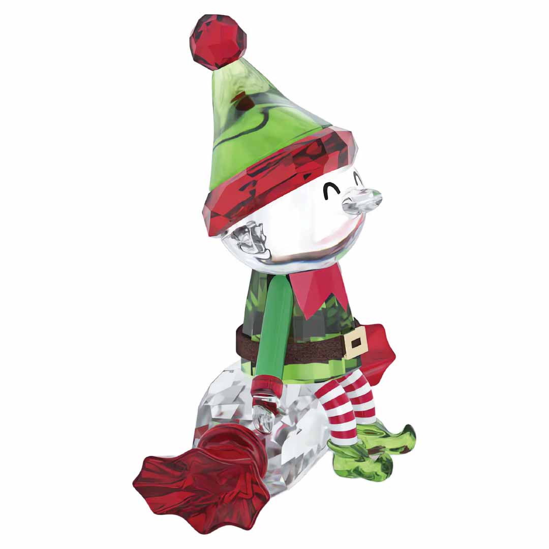 The Swarovski Holiday Cheers Dulcis Elf Home Décor piece embodies holiday charm with a cheerful elf dressed in a green outfit adorned with red accents. The elf, seated on a sparkling, faceted base, is detailed with red and white striped socks and has a red sack beside it, making it an enchanting festive decoration.