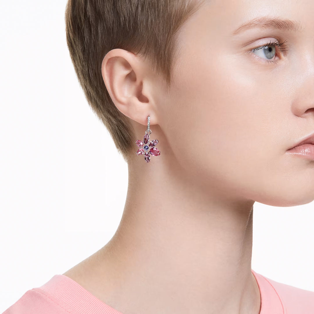 A person with short hair is wearing a pink top and elegant earrings, highlighted by the Swarovski Gema Drop Earrings from the renowned brand Swarovski. These mini hoop earrings feature pink gemstones in a floral design, enhanced with rhodium plating, against a plain white background.